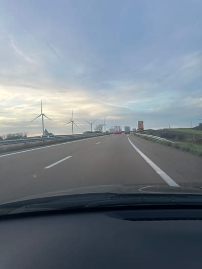The scene depicts a forward-facing view from inside a vehicle on a multi-lane highway, consistent with an autoroute near Sainte-Colombe, France. The lower quarter of the image displays the dark dashboard and windshield wipers of the observer's vehicle. The asphalt highway surface features distinct white dashed and solid lane markings, with at least two lanes visible in the direction of travel. Metal guardrails line both sides of the road.
On the elevated terrain to the left of the highway, a wind farm is visible, featuring at least five large, white-bladed wind turbines. In the mid-ground of the left lane, a white passenger vehicle is present. Further down the highway, a queue of vehicles, predominantly semi-trucks displaying illuminated red taillights, occupies both visible lanes, indicating a slowdown or stopped traffic. A large, rectangular road sign, yellow-orange in color with unreadable dark text/symbols, stands on the right shoulder beyond the congestion. The surrounding environment consists of rolling hills with green fields on the right and scattered trees and vegetation along the highway borders. The sky is overcast with varying cloud density, suggesting a cool, possibly early morning or late afternoon ambient light. No individuals are discernible.