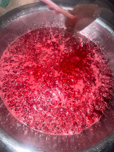 A close-up, overhead shot shows a large, shallow metal bowl filled with a bubbling mixture of mashed raspberries and their juices. The raspberries are a vibrant red, with some appearing whole and others broken down into a pulp. A light pink foam has formed on the surface of the mixture, indicating it is being cooked. A wooden spoon is partially visible on the left side of the bowl, its dark wood contrasting with the bright red of the fruit. The spoon is angled downwards, as if stirring the contents. The bowl itself appears to be made of stainless steel, with a slightly reflective surface that catches the light. The lighting suggests an indoor setting, possibly a kitchen, though no specific details of the surroundings are visible. The scene is focused entirely on the preparation of the raspberries, suggesting a process of making jam, jelly, or some other fruit preserve. The focus is sharp on the raspberry mixture, creating a sense of texture and depth. There is no visible text in the image. The overall impression is one of a rustic, home-style cooking process.
