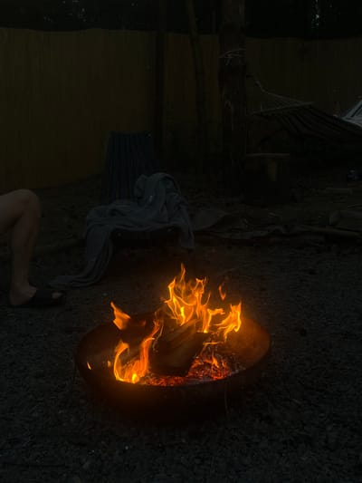 This image captures a nighttime scene in Ivachiv Horishnii, Ukraine, focused on a crackling campfire in a metal fire pit. The fire is intensely bright with vibrant orange and yellow flames licking upwards, illuminating the surrounding dark gravel ground and the charred edges of the pit. In the foreground, to the left, a person's lower leg and foot are visible, clad in a black sandal. Behind the fire pit, a dark blue wooden chair is draped with a light gray towel or blanket. Further in the background, a hammock with striped fabric is slung between trees, suggesting a relaxed outdoor setting. The environment appears to be a backyard or a campsite, enclosed by a wooden fence. The overall atmosphere is calm and rustic, characteristic of an evening spent outdoors.