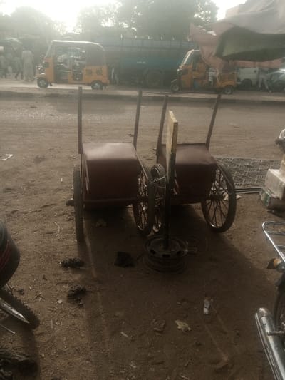 The image depicts a daytime street scene in Kofar Ruwa, Nigeria. In the immediate foreground, two dark red, two-wheeled handcarts with metal frames are positioned on unpaved dirt ground, oriented away from the camera. Between them, a vertical metal pole with an attached yellow rectangular sign is stabilized by a black metal car wheel rim at its base, with a small black tire and chain leaning against it. Partial views of two motorcycles flank the foreground: a front wheel and tire on the far left, and a chrome exhaust pipe and frame on the far right. The ground is dirt, containing scattered debris and dark clumps.
In the midground, a roadway runs horizontally, hosting vehicular and pedestrian traffic. Multiple yellow and black 'Keke Napep' auto-rickshaws are present; one prominent tricycle displays "TNA 4912" on its rear. A large blue flatbed truck is visible behind the tricycles. Pedestrians, some wearing light-colored traditional garments, are walking on the left side of the road. The background shows indistinct structures and sparse trees under a hazy sky.