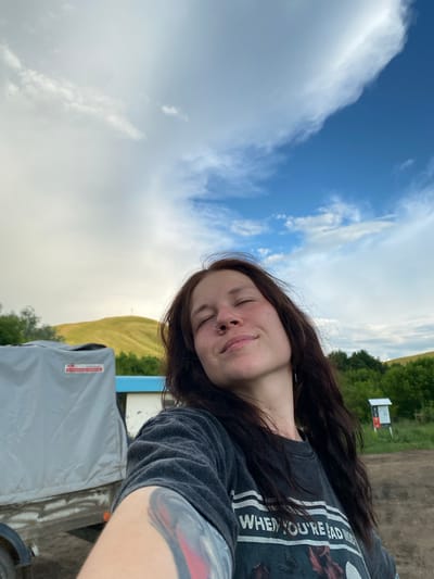 A young woman with long, dark hair and a nose ring takes a selfie outdoors. She is wearing a dark gray t-shirt with a graphic print. Her eyes are closed, and she has a relaxed smile on her face, suggesting a pleasant or contented emotion. Her arm, which is visible in the foreground, has a tattoo.

The background shows a vast sky with dramatic clouds, a mix of bright white and darker gray. Beyond the sky, there are rolling green hills and trees, indicating a rural or natural setting. A dirt road is visible in the upper right, and a signpost stands near the road, though the text on it is not legible. A light-colored tarp covers an object in the lower part of the image, with a red and white label visible on it. The overall impression is of an overcast day, possibly late afternoon, given the lighting. The scene appears to be located in Верхнеозёрное, Russia, suggesting an outdoor activity or a moment of personal reflection in a scenic location.