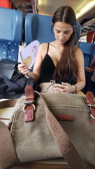 A young woman with long, dark hair is seated on a train, holding a purple fan in her left hand and a smartphone in her right. She is wearing a black sleeveless top and light-colored shorts. In front of her, a canvas messenger bag with leather accents is prominent in the foreground. The background shows blue train seats. The image is captured from a medium-low angle, focusing on the woman and the bag. The specific location within Považská Bystrica, Slovakia cannot be determined from the visual information provided.