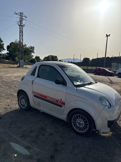 A white Abarth car is parked on a dirt and gravel lot under a bright, hazy sky. The car has a red and white Abarth decal on its side. In the background, a power pylon stands tall with wires stretching across the sky. Beyond the lot, there are trees, a fence, and a hint of mountains in the distance. The sun is high and creates a strong glare in the upper right portion of the image. The location is Vidreres, Spain.