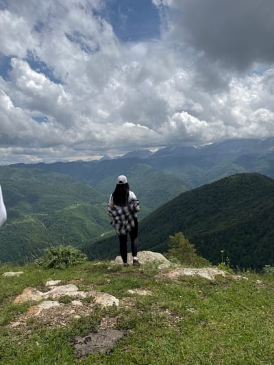 The image depicts a person standing on a rocky, grassy outcrop overlooking a vast, verdant mountain range. The person, viewed from behind, is wearing a white t-shirt, black leggings, a white baseball cap, and a plaid shirt draped over their shoulders. They are facing away from the camera, looking out at the expansive vista of densely forested mountains that recede into the distance, culminating in snow-capped peaks under a dramatic, cloudy sky. The location is identified as Nizhnyaya Teberda, Russia. The foreground consists of uneven terrain with rocks, sparse vegetation, and grass. The overall scene conveys a sense of awe and exploration amidst a majestic natural landscape.