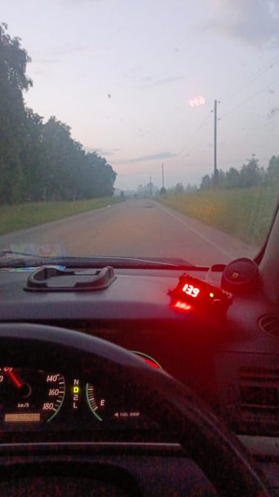 The image is taken from the interior of a car, looking out through the windshield at a road. The dashboard is visible in the foreground, with a steering wheel on the left. A digital display on the dashboard shows "139" in red. To the right of the dashboard, there is a small red digital device. The road ahead appears misty or foggy, with trees lining both sides. The sky is overcast. There are power lines visible in the distance. The location is identified as Zuy, Russia.