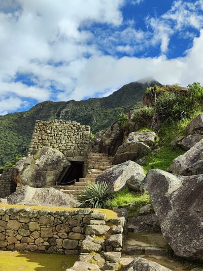 Ancient stone ruins nestle against a lush, green mountain backdrop under a sky with fluffy white clouds. A stone structure with a dark doorway stands beside stone steps that ascend upwards. Large, weathered boulders are scattered throughout the scene, interspersed with patches of grass and sparse vegetation. The stone structures exhibit intricate stonework and seem integrated with the surrounding natural elements.