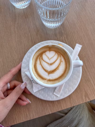 A close-up, top-down shot shows a white cup of coffee with latte art, sitting on a white saucer on a light brown wooden table. Two clear glass cups are visible in the upper portion of the frame. A person's hand with a ring and decorated fingernails is on the left side of the frame, gently holding the saucer. Two white sugar packets are positioned under the saucer. The coffee has a heart-shaped pattern created with milk foam. The scene appears to be in Donji Katun, Montenegro, likely at an outdoor cafe given the table setting and the natural lighting.