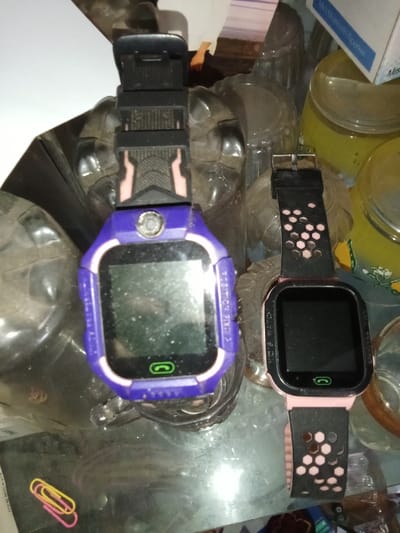 The scene is an indoor environment within the Emthanjeni Local Municipality, South Africa, featuring various objects resting on a reflective surface. Two smartwatches are the central subjects. The left smartwatch has a purple casing with a pink inner bezel, a black strap with pink accents, and a dark, dusty rectangular screen displaying a green call icon. A circular silver button is above the screen, and "T-TRITION PIX3" is embossed vertically on its right side. The right smartwatch has a black casing with a pink inner bezel and a black strap featuring hexagonal cutouts over pink material. Its rectangular screen is dark with a green call icon, and "YES WATCH" is embossed vertically on its left side. Two paper clips, one yellow and one pink, are visible in the lower-left. The background includes several translucent jars, some containing yellow/green substances, and a partially visible white and blue box labeled "Mini Bluetooth Speaker" in the upper right. The overall environment appears somewhat cluttered with uneven lighting.
