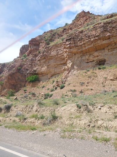 This image captures a rugged mountainous landscape in Getap, Armenia, under a blue sky with scattered white cumulus clouds. A diagonal pink-red streak, consistent with a camera lens flare or reflection, crosses the upper and middle portions of the frame from the top-left towards the mid-right.
The dominant feature is a large cliff face composed of stratified sedimentary rock, displaying varying hues of tan, brown, and reddish-brown. The rock surface is rough and uneven, with visible horizontal layering. Patches of sparse green vegetation, including small shrubs and grasses, are visible on the cliff's ledges and slopes, particularly on the upper ridge and at its base. A distinct, bright green bush is situated in a recess on the lower-mid section of the cliff face, towards the left. On the right side of the main rock formation, a dark, cave-like opening or overhang is present.
The foreground depicts a dirt and gravel roadside area, transitioning into a paved road fragment visible on the lower left, marked by a white lane demarcation line. The ground slopes gently upwards from the road towards the base of the cliff, covered with loose soil, small rocks, and scattered dry and green herbaceous plants. No individuals, animals, or artificial structures other than the road are visible within the scene.