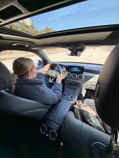 An interior shot of a black car, the sunroof open to reveal a clear blue sky above a tree line. A light-skinned woman with blonde hair in a braid is in the driver's seat wearing a gray patterned coat. She is holding her phone in her left hand and the steering wheel with her right. The car's dashboard, featuring the car's display and air vents, is visible. The car appears to be moving on a highway near Baltezers, Latvia.