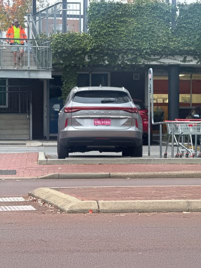 A photograph shows the rear of a silver Volvo SUV parked near a building in Bassendean, Australia. The license plate reads "GIRLBOSS" in pink letters. The car is parked close to the curb. A building with climbing greenery is visible to the left. In the background are shopping carts. A person wearing an orange safety vest stands by a metal scaffolding structure. The road surface is asphalt.
