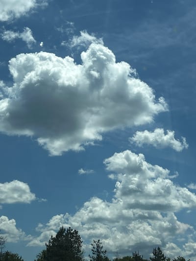 The image depicts a bright, partly cloudy sky over Verinsko, Bulgaria. Dominating the upper portion of the frame are large, fluffy cumulus clouds, some appearing bright white against the deep blue sky, while others show darker, shadowed undersides, suggesting depth and volume. The sunlight filters through and around these clouds, creating a bright and airy atmosphere. In the distance, a line of dark green trees forms the horizon beneath the sky. There are no people or any discernible activity in the scene. The image captures a serene moment of nature's grandeur.