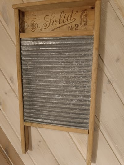 A vintage washboard hangs on a slatted wooden wall in Hovden, Norway. The washboard has a wooden frame and a corrugated metal washboard surface. The word "SOLID" is inscribed in large, decorative script across the top of the wooden frame, to the left of a circular emblem. Below "SOLID" and to the left of the emblem, the text "ASK RIKAT" is visible, along with a circular mark with what appears to be an abstract design. To the right of "SOLID," the number "Nr.2" is prominently displayed. The circular emblem to the right of "SOLID" contains more text and imagery, though it is difficult to decipher completely from this image. Within the emblem, there appear to be figures and the words "BONG," "BERGS," and "JUBILAE." The washboard is positioned vertically against the wall, with its top edge slightly above the halfway point of the frame. The wooden slats of the wall are light in color and run diagonally. The overall impression is one of a preserved antique in a rustic setting.