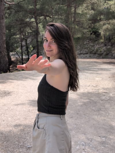 A young woman with long brown hair is captured from the waist up, facing away from the camera and looking over her shoulder with a slight smile. Her left hand is extended towards the viewer, palm open. She is wearing a dark, textured sleeveless top and light-colored pants. The background suggests an outdoor setting with trees and a dirt path, indicative of a natural landscape. The lighting is diffused, creating soft shadows. The image is taken at a medium distance, focusing on the woman and her immediate surroundings.