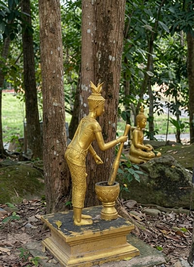 The image depicts two gold-colored statues in an outdoor, wooded setting. The foreground statue is of a standing figure with intricate detailing on its clothing and a headdress. This figure is positioned next to a mortar and pestle, with the pestle held in its right hand as if in the act of grinding. The statue stands on a tiered pedestal. In the background, to the right, a second gold-colored statue of a seated Buddha is visible, resting on a moss-covered rock. The environment appears to be a park or garden, with large trees, fallen leaves, and scattered rocks. The lighting suggests it is daytime. The image does not provide specific clues about its exact location within Sihanoukville, Cambodia.