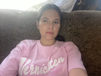 A young woman with dark hair and a nose ring is sitting on a couch. She is wearing a pink t-shirt with white text. The couch has a patterned fabric. Behind the couch, there is a window with sheer curtains and a decoration featuring a crescent moon and stars. The lighting suggests it is daytime, possibly indoors by a window. The image does not contain any information that would allow for the determination of the city of Romny, Ukraine, or any specific city.