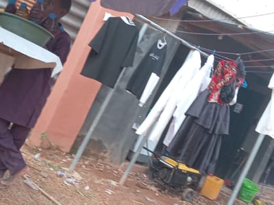 In Achara Ndiagu Omegio, Nigeria, a person carrying a tray with bottled drinks walks on a dirt ground. A clothesline holds various garments, including shirts, dresses, and other clothing items. A yellow gasoline generator and a container sit below the clothesline. A green basket is on the ground nearby.