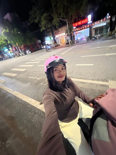 A young woman wearing a pink helmet and a brown long-sleeved shirt is riding a motorcycle at night on a wet street in Minh Xuân Ward, Vietnam. She is looking at the camera and smiling. The street is lit by streetlights and neon signs from businesses on the side of the road. There are other motorcycles and scooters parked on the side of the road, and some people are visible in the background. The woman is holding a pink bag.