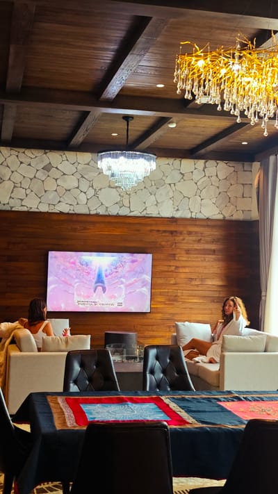 A medium shot shows the interior of a room, possibly in Cat Cat Village, Vietnam, as suggested by the prompt. The scene is dominated by a low-angle view featuring a wooden ceiling with beams and two chandeliers, one made of crystals and the other of a more modern design. The wall is a combination of stone and wood paneling. A flat-screen TV displaying colorful imagery hangs on the wooden portion of the wall. In the foreground, a table with a decorative blue, red, and gold cloth is surrounded by black chairs. On the opposite side of the table, two women are sitting on light-colored sofas. One woman, seen from the back, is wearing a white top. The other, facing forward, is wearing a white robe and pointing toward the camera. Between the women, a small cylindrical table is visible. The environment is luxurious, suggesting an upscale residence or hotel.