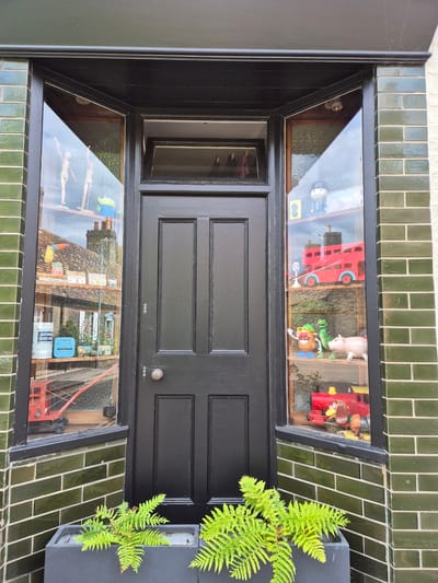 This is a forensic, matter-of-fact description of a shopfront in Cambridge, United Kingdom. The shop has a black wooden door with two panes. On either side of the door are bay windows. The shop is clad in dark green glazed bricks, with a light-colored brick base. In front of the shop are two dark grey planters containing bright green ferns.
The left window displays a variety of items. On a shelf, there are two wooden mannequins in a dancing pose and a green alien toy. Below this, there are some boxes and a blue sign. Further down, a red toy crane is visible.
The right window also displays toys. At the top, there is a toy figure in a black hat and suit. Below this, a red toy double-decker bus is displayed, along with a small white toy figure. Further down, there is a Mr. Potato Head toy, a green dinosaur toy, and a pink piggy bank. At the bottom, a red toy train is visible.
The reflections in the windows show buildings with tiled roofs and chimneys, suggesting a street view. The sky is visible in the upper parts of the reflections.