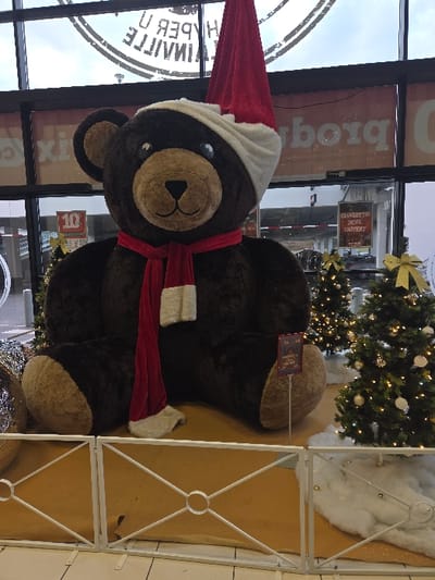 A large, dark brown plush teddy bear, wearing a red Santa hat with a white brim and a red scarf with white fringe, is centrally positioned in a festive display inside a commercial establishment. The bear sits upright, facing forward, with its lighter brown snout and paw pads visible. Behind the display, large glass windows reveal an outdoor parking lot under a cloudy sky. Reflected in the windows is the reversed text "HYPER U BLAINVILLE", indicating the location as a Hyper U supermarket in Blainville-sur-Orne, France. To the bear's right, two artificial Christmas trees decorated with warm white lights and gold bows are visible, set on a display floor covered with tan fabric and white faux snow. A low, white decorative metal fence with circular and cross patterns separates the display from the foreground. A small, dark red sign on a thin pole stands near the bear's right foot. To the bear's left, crumpled metallic decorations are present. No individuals are visible in the scene.