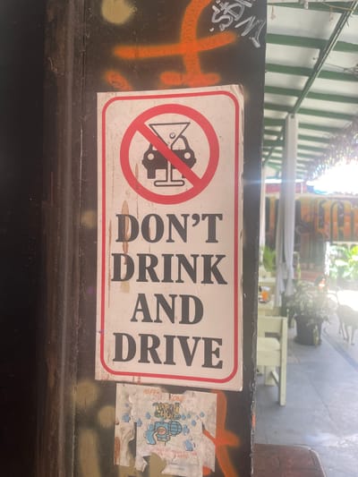 The image depicts a "Don't Drink and Drive" sign prominently displayed on a dark, textured surface, likely a wall or pillar, in Vagator, India. The sign itself is white with a red border and features a graphic of a car and a martini glass crossed out with a red circle, symbolizing prohibition. Beneath the graphic, the text "DON'T DRINK AND DRIVE" is clearly legible in large, bold, black capital letters. Below the main sign, there is a smaller, torn poster with colorful graphics and text, partially obscured. The background suggests an outdoor setting with elements of a cafe or restaurant, including tables, chairs, greenery, and a white awning. The lighting is bright, indicating daytime.
