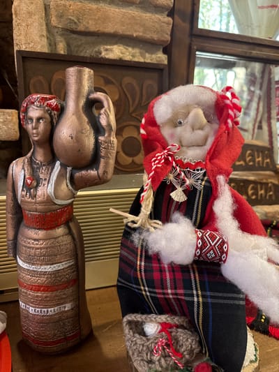 A still life photograph shows two objects. In the foreground, a bronze-colored statue depicts a person lying on their back, holding a ceramic jug on their stomach with both hands. The statue features red and white painted detailing on the head, chest, and body. In the background, a doll resembling an old woman wears a red and white striped hat and a plaid dress. It has white hair, round glasses, and a patterned band around its arm. The photograph was taken indoors in Kalimantsi, Bulgaria.