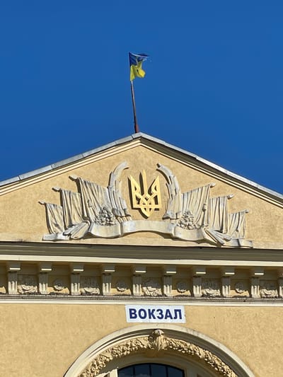The image shows the facade of a building with a Ukrainian flag flying from a flagpole on its roof. The building is painted a light yellow and features ornate architectural details, including a golden trident emblem and relief sculptures of draped flags and garlands. Below these decorations, a blue sign with white Cyrillic lettering reads "ВОКЗАЛ," which translates to "RAILWAY STATION." The building's design suggests it is likely an old, possibly historical, structure. The sky above is a clear, bright blue. This building is the railway station in the city of Stryi, Ukraine.