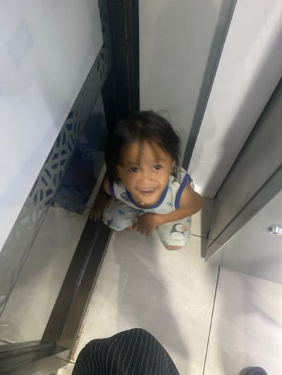 A young child, appearing to be a toddler, is crouched on a tiled floor indoors. The child is wearing a white sleeveless shirt with blue trim and a pattern of what appear to be sushi rolls or cartoon characters. The child's face is turned towards the camera, with a wide smile and dark hair falling across their forehead. The child's right hand is visible, resting on the floor.

The setting appears to be a modern interior, possibly a public space or a home with a contemporary design. The floor is tiled with light-colored square tiles. To the left and behind the child, there are metallic surfaces and white walls, suggesting a structure or fixture. The lighting indicates it is daytime, with diffused light casting soft shadows. The child's posture and expression convey happiness and a playful engagement with the camera. There is no visible text in the image. The overall scene is casual and focuses on the child's candid moment.