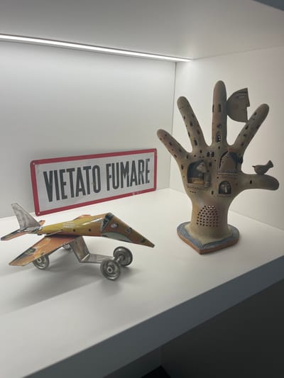 The scene, located in the city of Galatina, Italy, presents three static objects displayed on a white, flat shelf within a white-walled recess. An overhead linear LED light fixture illuminates the area. Positioned on the left, a multi-colored metallic toy airplane, predominantly orange and yellow with silver wings and tail, rests on four silver wheels, facing to the right. Behind the airplane and centrally located is a rectangular, white metallic sign with a red border, bearing the black uppercase inscription 'VIETATO FUMARE'. To the right, a ceramic sculpture depicts an upright, open human hand with an earthy, reddish-brown tone. The palm and fingers of the sculpture feature integrated miniature architectural elements, including windows and sculpted faces. A small bird figure is perched on the sculpture's outermost finger, and a separate facial element is partially integrated near the top of another finger. The sculpture stands on a base decorated with blue wavy patterns.