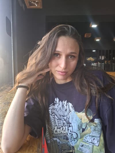 A woman with long brown hair is centered in the frame, looking at the camera. She's wearing a dark-colored t-shirt with a graphic design. Her left hand is near her face. A black hair tie is visible on her left wrist. The background includes dark wall and wooden surfaces, with what appear to be small, illuminated fixtures on the wall. Various objects are visible on a shelf in the background. The location is not identifiable as Sarandí, Argentina.