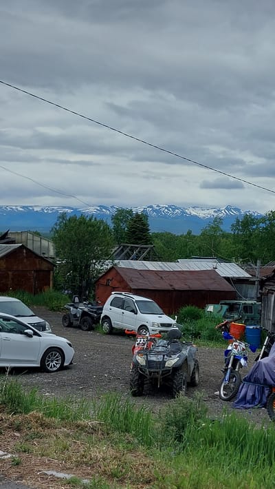 The image depicts a scene in an outdoor, rural area, possibly the city of Zelenyy, Russia, with a backdrop of snow-capped mountains. In the foreground, a gravel lot is occupied by several vehicles including a white sedan, a black ATV, a white compact SUV, a red ATV, and a dirt bike. There are also several dilapidated wooden structures, likely sheds or outbuildings, with rusty metal roofs. The vegetation consists of tall grass and some trees. The sky is overcast with gray clouds. The overall impression is one of a remote, perhaps slightly unkempt, residential or workshop area.