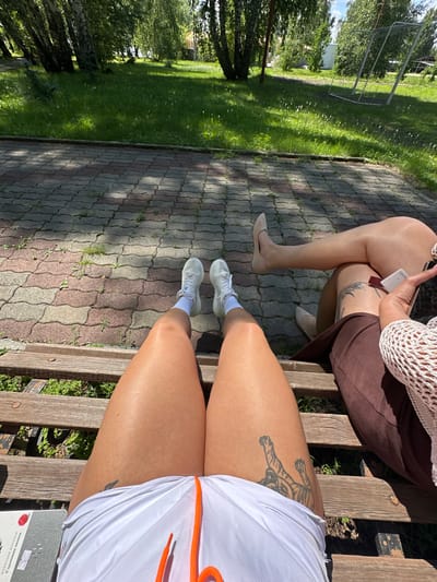 In Duvankul', Russia, two individuals are seated on a wooden bench in an outdoor setting, possibly a park or recreational area. The ground in front of the bench is paved with reddish-brown and gray interlocking bricks, partially covered with grass and weeds. Beyond the paved area, a grassy lawn stretches out, dotted with small white flowers. In the background, several trees with green foliage cast shadows, and a white building and a metal sports structure, resembling a soccer goal, are visible.

The person in the foreground is wearing white shorts with orange drawstrings and white sneakers with white socks. Their legs are bare, and a tattoo of a tiger is visible on their right thigh. They are holding a notebook or book with a red circular sticker on it.

The second person, seated to the right, is wearing light-colored pantyhose or sheer tights, a brown skirt, and light-colored pointed-toe shoes. They are holding a white card and a smartphone in their hands. A portion of a white, net-like garment is also visible. The sun is shining brightly, creating distinct shadows.