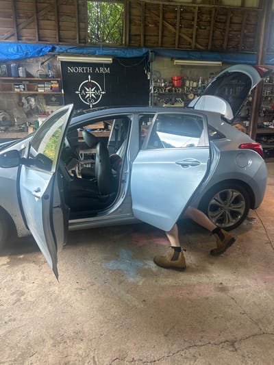 In a dimly lit, cluttered garage, a light blue hatchback car is positioned. The driver's side door and the rear hatch are open. A person's legs, wearing brown work boots and black socks, are visible reaching into the car from the driver's side. The garage environment shows shelves stocked with tools and supplies, a tarp draped from the ceiling, and a black wall with a white compass logo and text that reads "NORTH ARM CO". The floor is concrete and shows signs of wear and discoloration, with chalk drawings of a cross and what appears to be a butterfly. The overall scene suggests a person is working on or inside the vehicle within a workshop or storage space. The specific location in Thora, Australia is not determinable from the visual information alone.