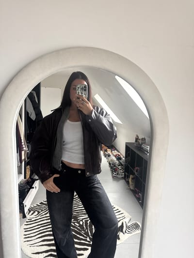 This image, reportedly taken in Tielrode, Belgium, depicts an indoor scene captured as a mirror selfie. A single individual, appearing female, is centered in an arched, off-white framed full-length mirror. The individual is wearing a dark brown leather bomber-style jacket over a white tank top and dark-wash straight-leg jeans. They are holding a smartphone, which features a silver-toned, patterned case, with both hands to capture their reflection. A ring is visible on the left ring finger.
The background visible in the mirror's reflection shows a room with white walls and a light-colored floor, partially covered by a white and black zebra-print rug. A sloped ceiling and an angled skylight window are visible in the upper right portion of the reflection. To the left of the individual, an open wardrobe or closet contains various hanging garments. To the right, a black modular shelving unit displays multiple pairs of sneakers and other items.