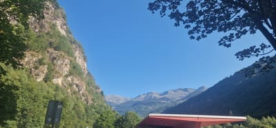 The image depicts a scenic mountain landscape in Lavorgo, Switzerland, under a clear blue sky. On the left, a steep, rocky mountain face is covered in dense green vegetation. In the foreground, a portion of a red roof structure is visible at the bottom right, partially obscuring the view of the valley and mountains beyond. Silhouetted tree branches with leaves frame the top of the image. The middle ground features a valley with smaller, tree-covered mountains and distant, rugged peaks. A grey traffic sign is present on the left side of the frame, with some text visible. The overall impression is of a natural, mountainous environment.