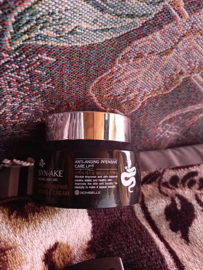 A dark-colored jar with a silver lid sits on a patterned fabric surface. The jar has text and a snake logo printed on it. The text on the jar reads "SYN-AKE NATURAL SKIN CARE INTENSE REPAIR WRINKLE CREAM" and "ANTI-AGING INTENSIVE CARE LINE". There is also Korean text on the jar and the brand name "BONIBELLE". The fabric appears to be a tapestry or blanket with a colorful, woven design. The lighting is soft, and the focus is on the jar. The image is a close-up shot of the product.