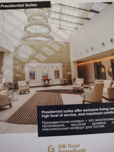 The image displays a spacious, modern hotel lobby with a high ceiling featuring a large, multi-tiered circular chandelier. The walls are a light neutral color, accented by geometric patterns and recessed circular lighting fixtures. Several plush armchairs and sofas are arranged in seating areas, with small tables between them. A striped rug covers a portion of the floor. In the background, artwork hangs on the walls, and doorways suggest further rooms or corridors. A black banner across the bottom of the image contains text in English and Russian, promoting "Presidential Suites" and describing their exclusive living, high level of service, and maximum comfort. Below the banner, the logo for "Silk Road Samarkand" is visible. The text in Russian appears to be a translation of the English description. There are no people visible in the image. The hotel is located in Bahshitpa, Uzbekistan.