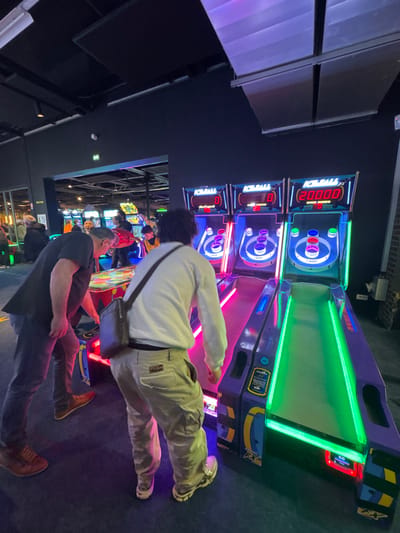 The image depicts an indoor entertainment venue, specifically an arcade, located in Saint-André-lez-Lille, France. In the foreground, two male individuals are actively engaged with arcade games.
On the right, a male subject is viewed from the back, wearing a white long-sleeved shirt, light-colored cargo pants, and white athletic shoes. He carries a dark shoulder bag diagonally across his torso. He stands in front of a row of three "ICE BALL FX" Skee-Ball style arcade games, which feature illuminated ramps and target holes. The game directly in front of him, and the one to its immediate left, display a score of "0" and "0 CREDITS." The leftmost "ICE BALL FX" game shows a score of "20000" and "0 CREDITS."
To the left of the primary subject, a second male is partially visible, seen from the torso down. He wears a dark short-sleeved shirt, dark pants, and brown shoes, leaning over a circular, multi-colored illuminated arcade game.
The environment is characterized by dark walls and a dark ceiling, featuring visible ventilation ducts and recessed lighting. The arcade floor is dark. Various other brightly lit arcade machines and indistinct figures of people are visible in the background, suggesting a busy atmosphere. An emergency exit sign is visible on the back wall. The lighting is primarily emitted from the arcade games themselves, creating a vibrant, low-light setting with strong blues, greens, and pinks emanating from the machines.