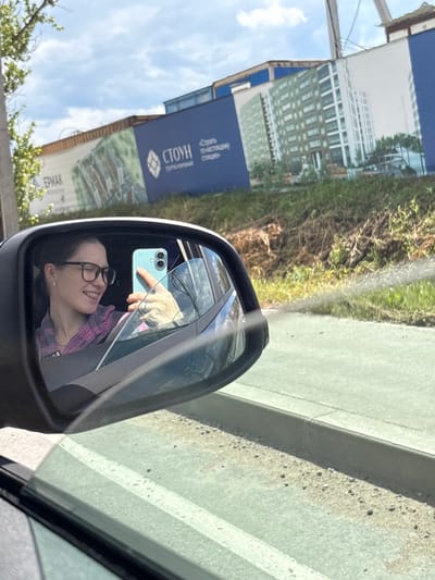 A woman is captured in a mirror selfie from inside a car, looking out a side window. She is wearing glasses and a pink and purple plaid shirt, with a ring on her left hand. The car's side mirror reflects her face, with her tongue sticking out playfully as she holds up a light blue smartphone to take the picture. The background visible in the mirror and through the car window shows a construction site with large billboards depicting apartment buildings and the text "СТОУН" and "ЕРМАК". The billboards are partially obscured by greenery and blue construction barriers. The scene takes place outdoors, under a partly cloudy sky, with what appears to be a road or parking lot in the foreground. The location is identified as Imeni Sverdlova, Russia.