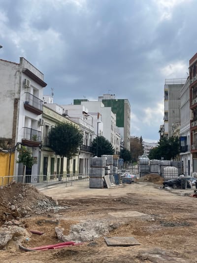 This image captures an urban street in Mérida, Spain, undergoing significant renovation or construction. The perspective is looking down a street or pathway, with buildings lining one side and a construction site on the other.
On the left side of the frame, a row of multi-story buildings stands under an overcast sky. These buildings display varied architectural styles; some feature traditional iron balconies and window shutters, while others are more modern with flat facades. Air conditioning units are visible on several buildings. Further down the street, a taller, slender building with a distinct green-paneled section on its upper stories rises above the other structures. Small trees and bushes are planted along the street in front of the buildings, with one tree near the foreground showing small, orange-like fruits.
The right side of the image is dominated by a rough, unpaved construction area, consisting of loose dirt, gravel, and some scattered debris. Several stacks of what appear to be paving stones or blocks are neatly arranged along the edge of this section, ready for installation. Large white cubic containers, likely IBCs (Intermediate Bulk Containers) possibly for liquids, are also present among the materials. Red pipes protrude from the ground in the foreground, and a metal grate or manhole cover is visible embedded in the dirt further back. A small, indistinct blue and white sign is visible among the construction materials.
The sky is heavily overcast with uniform grey clouds, indicating a cloudy or gloomy day. No people or vehicles are visible, suggesting the area might be closed off or the work is paused. The scene conveys a sense of an urban environment in transition, with ongoing infrastructure improvements.