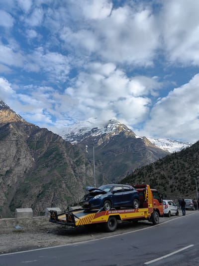 In the mountainous region of Kead, India, a tow truck is parked on the side of a road, with a damaged blue car loaded onto its flatbed. The blue car appears to have a crumpled hood and windshield, indicating it was involved in an accident. To the right of the tow truck, a white car is also parked, and several people are standing near it, some appearing to be onlookers or involved parties. The environment is characterized by steep, rocky mountains, some of which are snow-capped, and a cloudy sky with patches of blue. The road itself is paved, with a white line marking the lane. The scene suggests a traffic incident and the subsequent recovery process in a remote, mountainous area. The tow truck has "DEV BHOOMI RECOVERY SERVICES" written on its side.