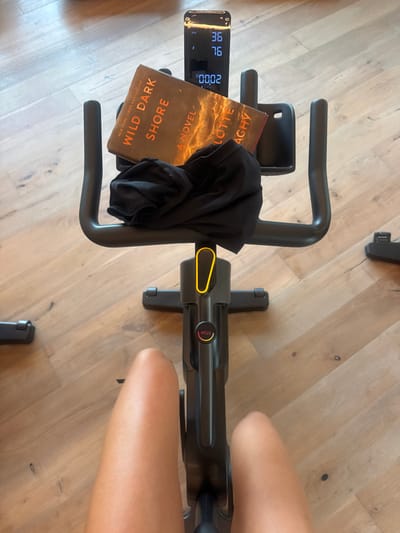 The scene depicts an individual seated on a black stationary exercise bike indoors in the city of El Caracol, Mexico. The perspective is a first-person view looking down. The subject's bare thighs are visible at the bottom of the frame, indicating a person is operating the bike. On the handlebars of the exercise bike, a hardcover book titled "WILD DARK SHORE A NOVEL BY CHARLOTTE MCCONAGHY" is resting, partially obscured by a crumpled black fabric item, likely clothing or a towel. A smartphone or digital display is mounted above the book, showing numeric values: "36," "76," and "0002." The bike features a yellow adjustment mechanism on its central column and a circular red "STOP" button with an orange outline below it. The exercise bike rests on a light-colored wooden floor with distinct grain patterns.