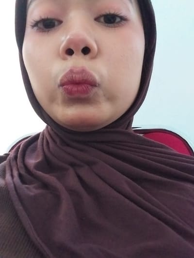 A close-up, forward-facing selfie shows a person wearing a dark purple hijab and top. The person's face is the central focus, with their lips pursed in a kiss. Their eyes are open, and their eyelashes are visible. The background is a plain, light blue wall, suggesting an indoor setting. The image was taken in Watampone, Indonesia.