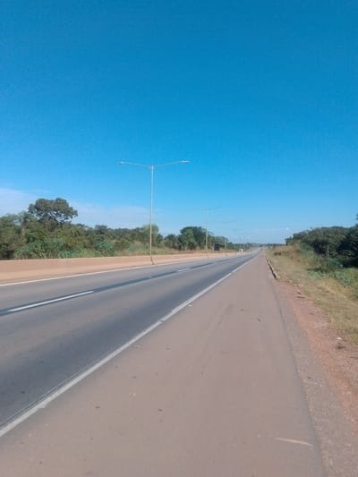 A photograph shows an empty, two-lane highway stretching into the distance under a clear blue sky. The highway is bordered by green trees and vegetation on both sides. Along the left side of the highway, a concrete barrier is visible. Several tall lampposts are spaced out along the median of the highway, indicating it is likely a highway designed for higher speeds or nighttime travel. The road surface appears to be asphalt and is in good condition. There are no vehicles or people visible in the image, suggesting a quiet and uninhabited stretch of road. The overall scene is one of a modern highway passing through a natural, rural landscape.