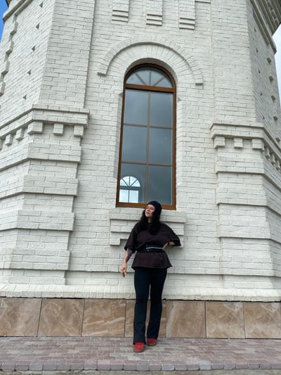 A full-shot, eye-level, outdoor photograph shows a young woman standing in front of a white brick building with an arched window. She is wearing a dark brown tunic with a black belt, black pants, a black beanie, and red sneakers. The woman is looking to her left with her right hand on her hip and her left arm bent at the elbow. The building appears to be made of white brick with decorative horizontal elements and a large, arched window with brown trim. The ground in front of the building is paved with red bricks and large, brown marbled tiles at the base of the building. The sky is overcast and the lighting is even. The location is specified as Andreevka, Russia.