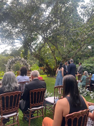 The image captures an outdoor formal event occurring in a lush garden environment within Camaragibe, Brazil. Dense green foliage, including mature trees with visible epiphytes, forms the backdrop, and sunlight penetrates through the canopy. The ground is covered in short green grass.
In the foreground, the backs of three individuals are visible, seated in dark wooden Chiavari-style chairs with white cushions. From left to right, these include a person with long, wavy gray hair in dark clothing, a person with short gray hair in a dark suit, and a person with long dark hair wearing a black sleeveless top and orange pants. White flowers are scattered on the grass between the chairs.
In the mid-ground, a central group of four individuals stands. This group comprises a woman in a light blue dress with shoulder straps, flanked by three men. Two men are dressed in dark suits, facing the woman and each other, appearing engaged in a conversation or ceremony. A third man, wearing a light brown suit, stands to the right of this group, observing. Other seated attendees in the mid-ground include a person with curly dark hair wearing a striped shirt and an older woman with light hair in a light green dress, both observing the central standing group. To the far left in the mid-ground, a decorative arch adorned with white flowers is visible, positioned over a set of empty chairs. Overhead, string lights are suspended between the trees. The overall scene indicates a formal gathering, likely a wedding ceremony or similar celebration.