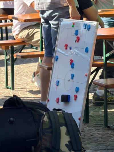 The image depicts a whiteboard set up on a table at an outdoor seating area, likely at a restaurant or cafe. The whiteboard displays a diagram representing a soccer or hockey game formation, with blue and red magnetic pieces arranged to represent players. A black eraser is also visible on the board. Several individuals are seated around the table, indicated by their visible clothing, including denim shorts, khaki pants, and a white shirt. A backpack, partially obscured in the foreground, has a camouflage print on its upper portion and is black on the lower portion. The location of the scene is Fentange, Luxembourg.