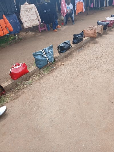 The image captures an outdoor market scene in Hospital ward, Kenya, characterized by an unpaved, light brown granular surface extending from the foreground to the midground. A line of seven handbags is displayed along a low, raised concrete curb bordering a narrow strip of dry vegetation. From left to right, these bags include a red top-handle bag with white trim, a large teal tote bag, a black duffel bag, a black structured shoulder bag, two light brown structured handbags with metallic hardware, and a dark red shoulder bag. In the background, various garments are suspended on what appear to be makeshift display racks, featuring items such as a black puffer jacket, an orange puffer jacket, a blue jacket, a beige puffer jacket with "SWEET" emblazoned on it, and other assorted clothing. Three individuals are partially visible in the background: one person stands in a white t-shirt and dark trousers, another's lower body is visible wearing blue jeans and orange shoes while seated on a purple stool, and a third individual is partially obscured. A foot wearing a white athletic shoe is visible in the lower-left foreground. The scene is illuminated by natural daylight.