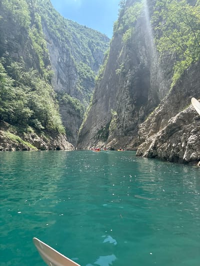 The image depicts a group of kayakers navigating a narrow gorge with towering, verdant cliffs on either side. The water is a striking turquoise color, calm and reflecting the sunlight. The sun is shining brightly, creating a dappled light effect on the rock faces. Dense green vegetation clings to the steep slopes, adding to the wild, natural beauty of the scene. In the distance, several kayaks are visible, forming a small procession. The perspective is from one of the kayaks, with a paddle blade partially visible in the foreground. The overall impression is one of peaceful exploration and immersion in a dramatic natural landscape. The time of day appears to be late morning or early afternoon, given the strong sunlight and the angle of the shadows. The weather seems to be fair and warm. The location is suggested to be Dubrovsko, Montenegro, a region known for its stunning natural gorges.